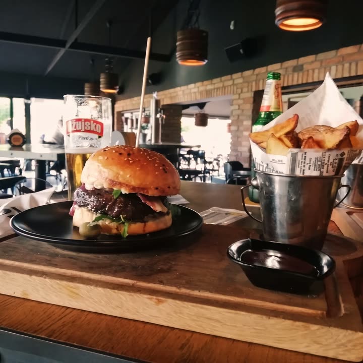 Burger s roštilja na crnom tanjuru, uz čašu točenog Ožujskog i kanticu pečenog krumpira